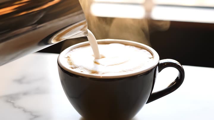 A barista pours steamed milk into a flat white on a marble c