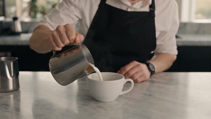 A barista pours steamed milk into a flat white on a marble c